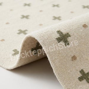 May include: Close-up of a cream-colored rug with a repeating pattern of olive green cross shapes and small brown dots. The rug's edge is folded over, revealing its woven texture. The rug is made of a thick, durable material.