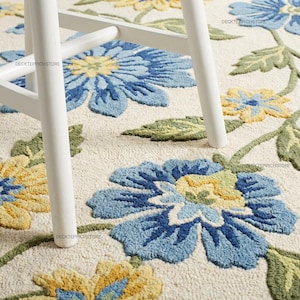 May include: A tufted area rug featuring a cream-colored background with a repeating floral pattern in shades of blue, yellow, and green. The rug is partially covered by the white wooden legs of a piece of furniture, with the text DECKTEPPICHSTORE visible.