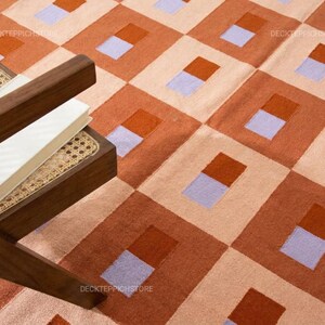 Puede incluir: Una alfombra con un patrón de cuadrícula geométrico repetitivo en tonos terracota, melocotón claro y lavanda. Cada cuadrado contiene un bloque rectangular más pequeño dividido en dos colores. El texto DECKTEPPICHSTORE es visible en la alfombra.
