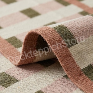 May include: A woven rug featuring a repeating checkered pattern in muted olive green, soft dusty pink, and cream. The fabric has a visible textured weave and is folded over to reveal the underside, with the text deckteppichstore visible.