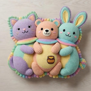 May include: A handmade crochet pillow featuring a cat, bear, and bunny. The plush toys are in pastel colors, including lavender, peach, and mint green, with rainbow accents. The bear has a honey pot applique. The pillow is designed for decorative use.