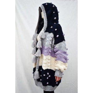 Puede incluir: Un suéter con capucha de gran tamaño hecho a mano, con un diseño de patchwork. La capucha azul marino tiene pompones y borlas grises. El cuerpo del suéter tiene rayas grises, lavanda, crema y azul marino. Las mangas son largas y el ajuste es holgado.