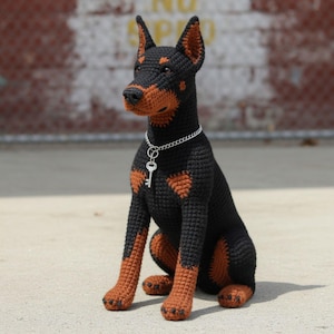 May include: A handmade crochet Doberman Pinscher dog in black and tan. The dog is sitting with a silver chain necklace and key pendant. The dog's ears are upright, and it has a detailed snout and paws.