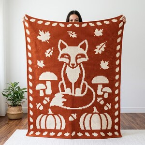 May include: A rectangular crochet blanket featuring a burnt orange background with a cream-colored pattern. The design displays a central fox, mushrooms, acorns, and pumpkins, all framed by a border of small leaf shapes.