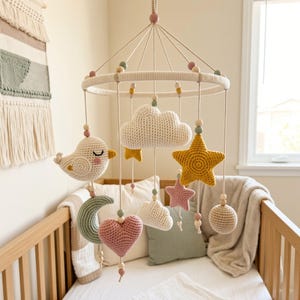 May include: A hanging nursery mobile featuring crocheted figures including a white bird, a large white cloud, a yellow star, a pink star, a sage green crescent moon, a pink heart, and a beige sphere, all suspended from a circular frame with wooden beads.