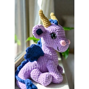 May include: A handmade crochet dragon in shades of purple, blue, and gold. The plush toy features large, expressive eyes, a pink snout, and detailed wings. The dragon is sitting on a windowsill, showcasing its intricate design and craftsmanship.