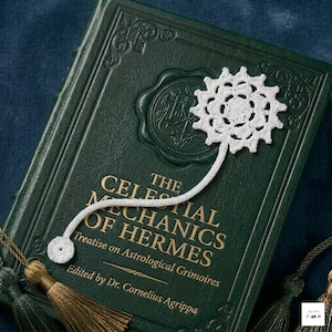 May include: A white crocheted bookmark with a circular floral motif and a small round end piece, resting on a dark green hardcover book titled The Celestial Mechanics of Hermes, Treatise on Astrological Grimoires, Edited by Dr. Cornelius Agrippa.