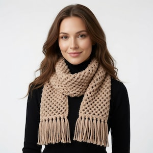 Könnte beinhalten: Ein beigefarbener Häkelschal mit einem durchbrochenen Gittermuster und langen Fransen an beiden Enden. Der Schal wird über einem schwarzen Langarmshirt getragen. Die Strickstruktur ist durchgehend sichtbar und zeigt ein gleichmäßiges Garn.