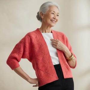 Peut inclure: Un cardigan corail à devant ouvert avec un motif géométrique en losanges. Les manches trois-quarts et la coupe décontractée créent une silhouette confortable. Le cardigan est associé à un haut blanc et un pantalon noir. Un bracelet doré est visible.
