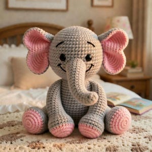 Peut inclure: Un jouet éléphant crocheté à la main, de couleur grise avec des détails roses. L'éléphant a de grandes oreilles doublées de rose, une trompe et un visage souriant avec des yeux noirs. Les pieds et l'intérieur des oreilles sont roses. Le jouet est posé sur un lit.