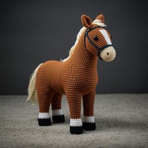 May include: A handcrafted crochet horse toy in shades of brown, cream, and white. The horse features a white blaze on its face, a cream-colored mane and tail, and black hooves with white accents. It is wearing a blue bridle.