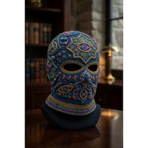 May include: A blue, orange, and purple crocheted balaclava with an intricate design. The mask-like head covering features floral and geometric patterns, with eye and nose openings. The neckband has stripes of green, orange, and purple. The balaclava is displayed on a black mannequin head.