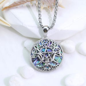 Puede incluir: Un collar con colgante circular de tono plateado que presenta un intrincado diseño de árbol de la vida entrelazado con un pentagrama central y símbolos de media luna. El colgante tiene una incrustación de concha de abulón iridiscente.