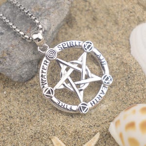 May include: Silver-toned pendant necklace featuring a pentagram within a circular frame. The frame is inscribed with the words "Spirit", "Air", "Earth", "Fire", and "Water". The necklace rests on sand with seashells and a rock in the background.