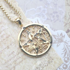 May include: A gold-toned circular pendant necklace featuring an intricate openwork pentagram design. The metal surface is engraved with various symbols, Hebrew characters, and the word GRAMMATON along the outer rim, suspended on a box chain.