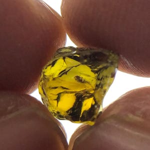 May include: A close-up of a rough, uncut gemstone held between two fingers. The gem is a vibrant yellow color with dark inclusions and a slightly translucent appearance. The gem's surface has a textured, irregular shape.