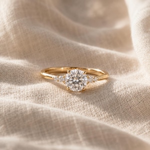 Può includere: Un anello in oro con una grande pietra centrale rotonda a taglio brillante, affiancata da tre pietre rotonde più piccole su ciascun lato della fascia. La fascia in metallo è in oro giallo lucido e le pietre sono chiare e sfaccettate.
