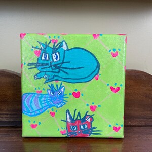 Könnte beinhalten: Quadratische Leinwandkunst mit drei Cartoon-Katzen in Türkis, Blau und Rot. Die Katzen sind vor einem limettengrünen Hintergrund mit einem rosa Herz- und Rautenmuster platziert. Das Kunstwerk ist eine skurrile und farbenfrohe Darstellung von Katzen.