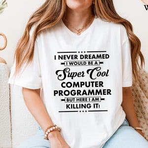 Op de afbeelding: Wit T-shirt met de tekst "I NEVER DREAMED I WOULD BE A Super Cool COMPUTER PROGRAMMER BUT HERE I AM KILLING IT!". Het shirt heeft een ronde hals en korte mouwen. De tekst is zwart.