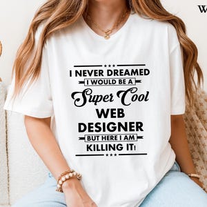 Puede incluir: Camiseta blanca con el texto "I NEVER DREAMED I WOULD BE A Super Cool WEB DESIGNER BUT HERE I AM KILLING IT!". El texto es negro, con una mezcla de fuentes y estilos. Una prenda informal.