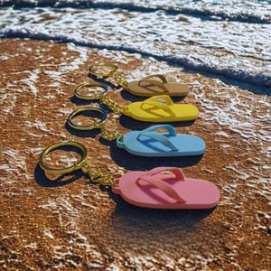 May include: Four colorful flip-flop keychains in brown, yellow, blue, and pink, with gold key rings and chains, resting on a sandy beach. The keychains are arranged in a row, with the ocean visible in the background.