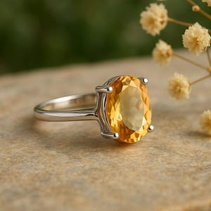 May include: A silver ring featuring a large, oval-cut citrine gemstone. The bright yellow-orange stone is set in a four-prong setting, with a simple, polished silver band. The ring is displayed on a neutral-toned surface.