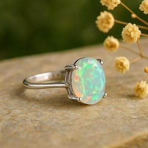 Può includere: Un anello d'argento con un'opale ovale. L'opale mostra colori iridescenti di verde, blu e rosa. L'anello è posto su una superficie neutra con uno sfondo sfocato e rametti di fiori gialli.
