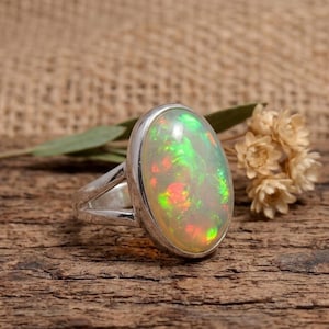 Könnte beinhalten: Ein Silberring mit einem großen, ovalen Opal-Edelstein. Der Opal zeigt ein lebendiges Farbspiel mit grünen, roten und gelben Blitzen auf einer milchig-weißen Basis. Der Ring liegt auf einer rustikalen Holzoberfläche.