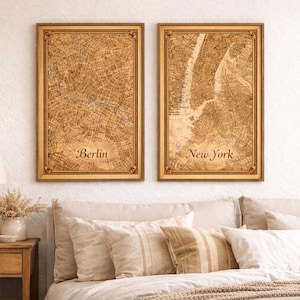 Op de afbeelding: Twee ingelijste stadskaarten, één met het label "Berlijn" en de andere "New York", hangen boven een bed. De kaarten hebben een vintage esthetiek met een lichtbruin kleurenschema. De frames zijn goudkleurig. Het bed heeft beige kussens en een crèmekleurig dekbed.