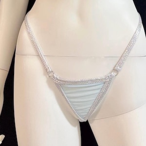 Puede incluir: Ropa interior de estilo tanga azul claro con ribete y tirantes de purpurina plateada. La parte delantera triangular es azul claro y las correas son finas y brillantes. La ropa interior se exhibe en un maniquí.
