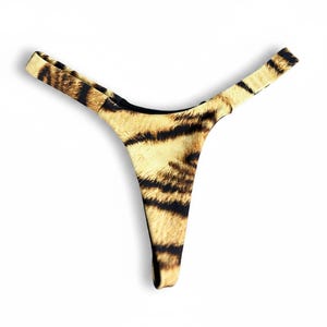 Puede incluir: Una braguita de estilo tanga con un diseño de estampado de tigre. La prenda presenta una pernera de corte alto y una cobertura trasera mínima. La tela tiene un patrón amarillo y marrón, que imita el aspecto de la piel de tigre. El tanga está diseñado para un ajuste cómodo.