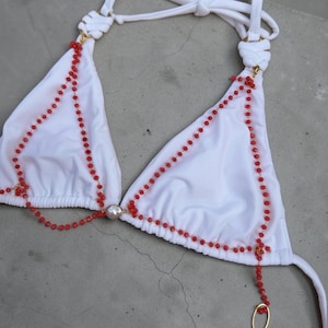 Puede incluir: Top de bikini blanco con detalles de cuentas rojas. El top triangular presenta cuentas rojas que delinean los bordes y forman una cadena decorativa. Una gran cuenta con forma de perla está en el centro. Las correas son blancas.