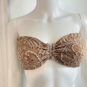 Puede incluir: Un top tipo bandeau sin tirantes con una capa base de color carne cubierta por un encaje beige intrincado. El encaje está adornado con pequeñas lentejuelas a juego. Las dos copas están unidas en el centro por un pequeño cierre de anillo metálico plateado.