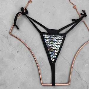 Puede incluir: Braga de tanga negra con un panel frontal triangular cubierto de formas geométricas iridiscentes. La prenda tiene tirantes finos y se muestra en una percha de color cobre sobre un fondo gris. Un artículo de lencería.