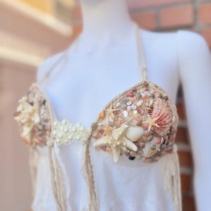 Può includere: Un top all'americana bianco con un corpetto decorato con varie conchiglie, stelle marine e piccole perle bianche. Il capo presenta spalline intrecciate e dettagli con frange pendenti, creando una superficie testurizzata sulle coppe.