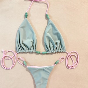 May include: A two-piece bikini set in a soft, muted green color. The bikini top and bottom are connected with pink string ties and adorned with green beads. The bikini top has triangle cups and the bottom is a thong style.
