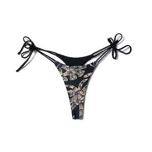 Puede incluir: Braguita de bikini negra y estampada con laterales anudados. La braguita presenta un fondo oscuro con un diseño de hojas tropicales beige. Las cintas son negras y la braguita del bikini es de estilo tanga.