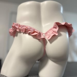 May include: A light pink thong featuring a ruffled trim along the waistband. The garment is displayed on a white mannequin torso, showcasing the gathered fabric detail and the thin strap design that connects at the center back of the piece.