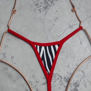 Puede incluir: Un tanga de bikini con estampado de cebra rojo y negro. La parte inferior del bikini está colgada de una percha de color cobre sobre un fondo gris. El tanga tiene un ribete rojo y un estampado de cebra.