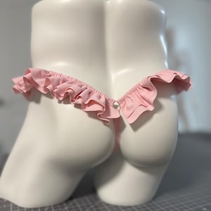 May include: A light pink thong featuring a ruffled trim along the waistband. The garment is displayed on a white mannequin torso, showcasing the gathered fabric detail and the thin vertical strap connecting the back section to the waistband.