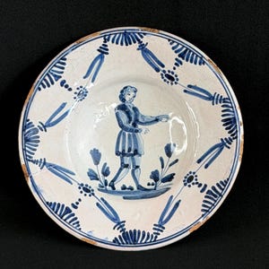 May include: Antique white ceramic plate with a blue and white design. The plate features a central figure in blue attire, surrounded by floral and ribbon-like patterns. The rim has a decorative border. The plate shows signs of age and wear.