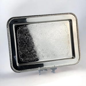 Op de afbeelding: Een rechthoekige zilveren serveerschaal met een decoratief bloemmotief en een touwachtige rand. De schaal wordt ondersteund door een kleine, transparante standaard. Het oppervlak reflecteert licht.