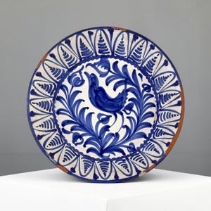 Op de afbeelding: Een ronde keramische bord met een handgeschilderd blauw vogelmotief, omringd door complexe blauwe bloem- en bladpatronen op een witte achtergrond. De buitenrand toont een herhalend waaiervormig ontwerp en de rand heeft een terracotta kleur.