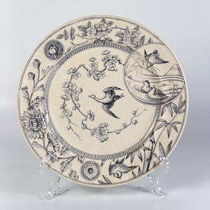 Puede incluir: Un plato redondo, blanquecino, con un diseño detallado en azul y blanco. El plato presenta motivos florales y de aves, incluyendo patos volando y patos en un estanque. El borde tiene un patrón geométrico y detalles florales.