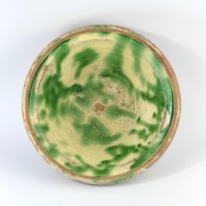 May include: A round, antique ceramic bowl with a cream-colored interior and a green, mottled pattern. The rim is a reddish-brown color. The bowl has a slightly uneven surface, suggesting it is handmade. The bowl is likely for serving food.
