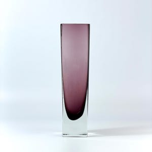 Könnte beinhalten: Eine hohe, rechteckige Glasvase mit einem Farbverlauf von Lila zu Klar. Die Vase hat einen quadratischen Boden und einen abgerundeten Innenboden. Das Glas ist glatt und reflektiert das Licht, wodurch eine moderne Ästhetik entsteht.