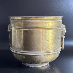 Peut inclure: Un pot en laiton vintage avec un corps rond et un large bord. Le pot comporte deux poignées décoratives avec des visages sculptés. Le laiton a une patine chaude et vieillie, lui donnant un aspect classique et antique.