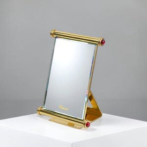 May include: A rectangular table mirror with a polished gold-tone metal frame. The frame features cylindrical top and bottom bars, each accented with a round red cabochon at the ends. The word Chopard is printed in gold on the lower mirror surface.