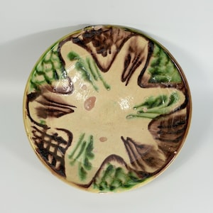 May include: A round ceramic bowl featuring a cream-colored base with a hand-painted design of dark brown and green abstract shapes. The interior surface displays a glossy finish with visible brushstrokes and a repeating pattern of dark brown lines.