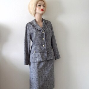 Puede incluir: Un traje de falda de tweed gris y blanco con cierre de botones blancos y un bolsillo en la parte delantera. El traje lo lleva un maniqu&iacute; con un sombrero de piel blanca.